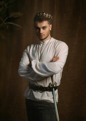 Portrait of handsome man in image of medieval king. Vintage clothing, historic white shirt retro clothing gold crown on head. Gothic room, studio. Fashion model posing. Adult strong sexy guy knight.