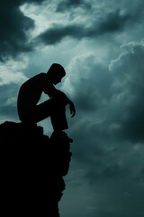 Silhouette of a man sitting alone on a cliff edge under dark, dramatic clouds. Concept of sadness, depression, and loneliness.