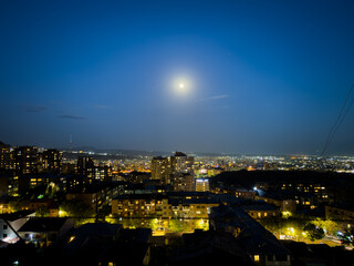 The city is lit up at night with a full moon shining brightly in the sky. The moon is positioned in the middle of the sky, casting a soft glow over the city below