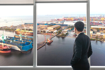 Businessman in suit looking out office window at shipping port, symbolizing logistics, trade, and...