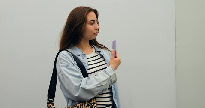 Fashionable young woman with a denim jacket and leopard print bag takes selfies with her purple smartphone, trying various poses against a minimalist gray background. concept of shopaholism in women