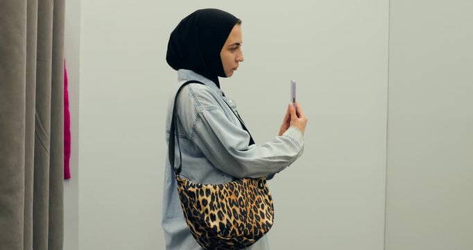 Fashionable muslim woman wearing hijab and denim jacket takes selfies with smartphone in dressing room mirror, adjusting pose and expression. concept of freedom for Muslim women