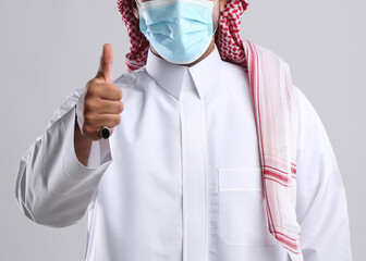 saudi arabian man in thob standing wearing face mask front facing showing thumb finger