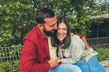 Happy couple laughs as boyfriend tickles girlfriend on park bench. Joyful moment of affection,...