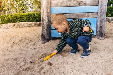 2 year old boy plays in sandbox holding a yellow shovel. He has a pacifier in his mouth and enjoys...