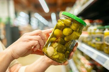 Consumer woman reading label on glass jar of pickles before buying in supermarket close-up.