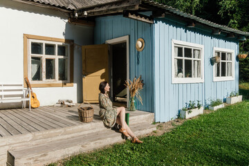 A woman enjoys a peaceful moment sitting on the steps of a blue cottage with a wooden porch....