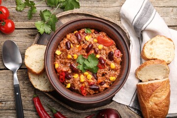 Tasty chili con carne in bowl, ingredients and bread on wooden table, flat lay