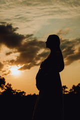 Fototapeta premium beautiful silhouette of pregnant woman with long hair at sunset , clouds and sky. third trimester of pregnancy, expecting baby, health of expectant mother and child. walk on summer day. shadows