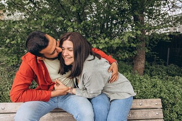 Happy couple laughs as boyfriend tickles girlfriend on park bench. Joyful moment of affection,...