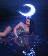 Fantasy woman queen of night lady moon, crown glows on head, black dress cape shining stars fairy girl goddess creative costume beauty face. Summer dark nature water lake river sky sunset art portrait