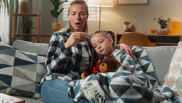 Caucasian family at home medicine health pandemic mother cuddling with daughter checking temperature with thermometer high fever unhealthy sick kid girl mom woman caring child flu childcare concept - Powered by Adobe