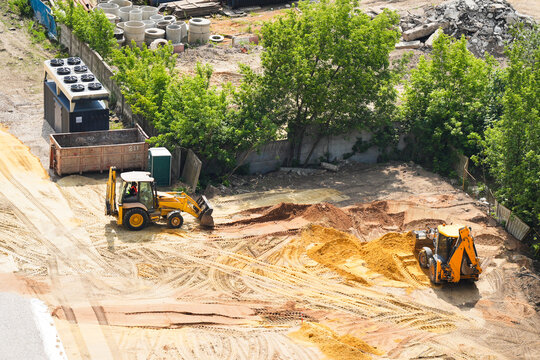 Two yellow excavators working on a construction site, moving sand and soil near trees and heavy equipment. Aerial top-down photo of groundwork, earthmoving and site preparation for development