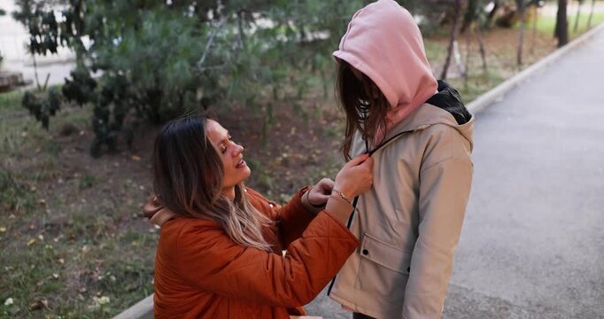 Loving mother zips hooded coat of little daughter in city park. Blonde woman cares of preteen girl walking together about autumn town slow motion