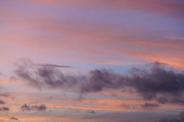 Vibrance of fiery orange clouds crossing over a light blue evening sky is suppressed by wafting puffs of low flying clouds showing darkened pastel hues of violet and subtle red mixed into gray base.