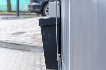 A black waste bin securely mounted on a wall next to a paved walkway in a city area, surrounded by...