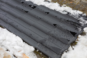 Black roof panels lie on the ground, coated with snow and water droplets, highlighting harsh winter...