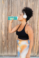 Curvy Afro woman drinking water after exercising outdoors, wearing headphones and sportswear
