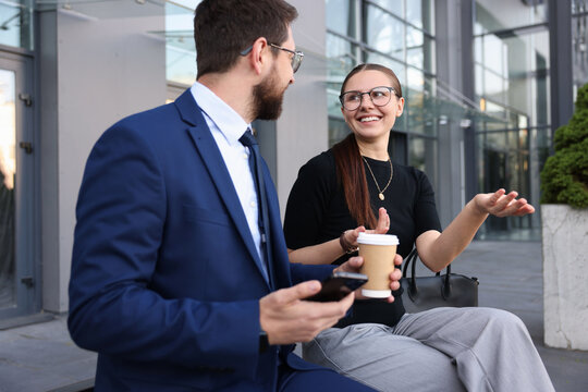 Happy coworkers with smartphone and coffee discussing something near office building