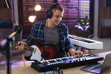 Smiling man in headphones working with MIDI keyboard and guitar at table in home studio