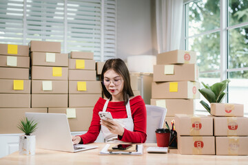 Young Asian woman in red shirt and apron doing online business, multitasking between smartphone and...