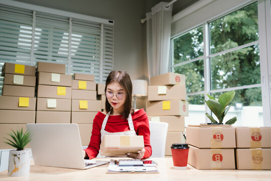 Young Asian woman in red shirt and apron doing online business, multitasking between smartphone and laptop, surrounded by packaging boxes in modern workspace.