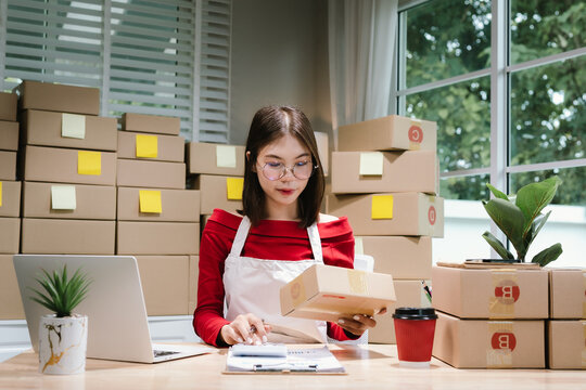 Young Asian woman in red shirt and apron running her online business, multitasking between smartphone and laptop, surrounded by packaging boxes in modern workspace.