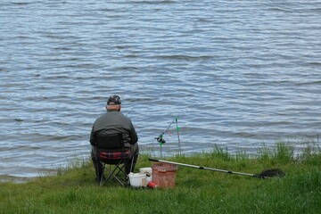 Sitting old man is fishing with fishing rod on coast of lake.