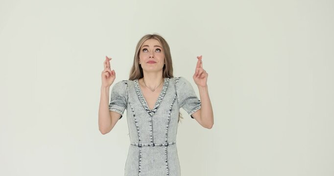 Woman with long light hair in denim dress holds fingers crossed praying. Crossed fingers suggest hoping for good luck sign and wish to come true