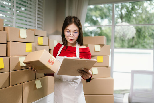 Young Asian woman in red shirt and apron doing online business, multitasking between smartphone and laptop, surrounded by packaging boxes in modern workspace.