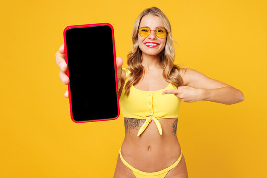 Young sexy woman wear bikini swimsuit rest near hotel pool hold in hand use point on mobile cell phone with blank screen area isolated on plain yellow background. Summer vacation sea sun tan concept. - Powered by Adobe