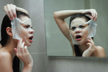 Surprised young woman applying facial mask in front of mirror, expressing curiosity and concern...