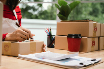  A hardworking woman in a red shirt and apron manages online orders, multitasking between packaging boxes and using her laptop in a busy e-commerce and shipping workspace.