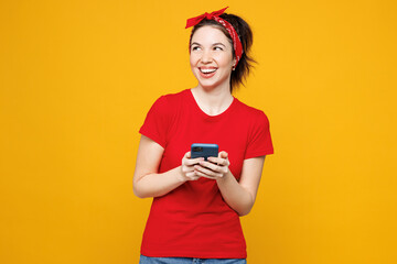 Young smiling happy woman she wearing red t-shirt casual clothes hold in hand use mobile cell phone looking aside on area mockup isolated on plain yellow background studio portrait. Lifestyle concept.