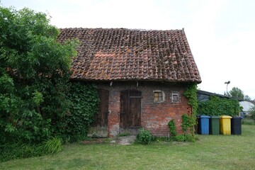 Old brick farm building with red roof tiles and wooden doors in countryside. Colorful waste sorting containers next to building.