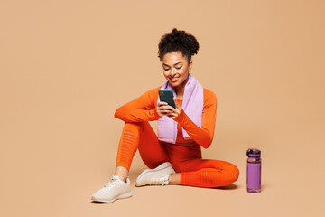 Full body young fitness trainer instructor sporty woman sportsman in orange clothes spend time in gym sit hold use mobile cell phone isolated on plain beige background. Workout sport fit abs concept