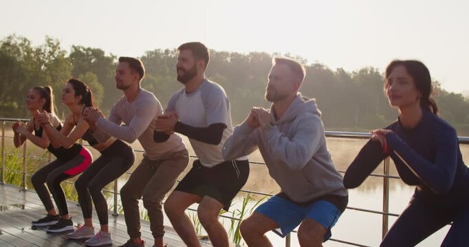 Sporty smiling happy group people doing sport sits up exercises in nature at sunset. Young men and women group in sportswear having workout in the park. Outdoors training and healthy concept.