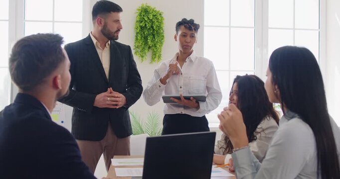 Business people in formal clothes clapping hands greeting new male employee sitting om workplace in office. Young African American woman introducing new coworker standing in meeting room.