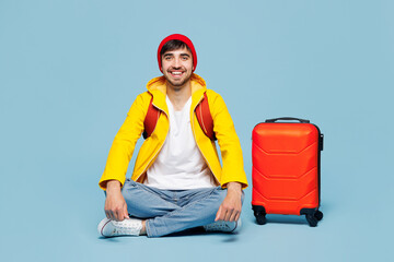 Traveler smiling happy man wear hat yellow casual clothes sit near bag isolated on plain pastel blue background. Tourist travel abroad in free spare time rest getaway. Air flight trip journey concept.