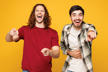 Young two friends buddies men they wearing casual clothes together pointing finger on camera with smile mocking or humiliate isolated on plain yellow orange color background studio. Lifestyle concept.