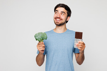 Young man he wear blue t-shirt casual clothes hold choose between chocolate bar and broccoli look...