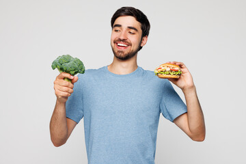 Young smiling man wear blue t-shirt casual clothes hold broccoli choose between vegetables and...