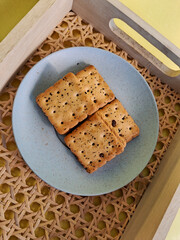 Square crackers with black sesame seeds served on a minimalist blue plate, placed in a woven wooden...