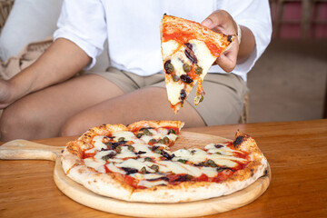 Hand lifting hot cheesy pizza slice with toppings, rustic wood table, casual dining vibe, perfect...