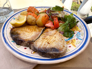 Grilled tuna fish steak served on a decorative patterned plate with fresh vegetables, lemon wedges, and boiled potatoes, showcasing a delicious Canarian traditional culinary presentation