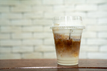 Iced coffee with creamy milk layers in a clear plastic cup, topped with frothy whipped cream and sprinkled with cinnamon. Placed on a wooden surface with a light brick background.