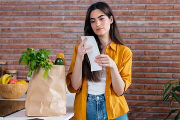 Beautiful woman checking shopping ticket while leaving cardboard bag with healthy shopping in...