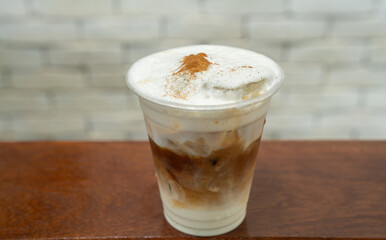 Iced coffee with creamy milk layers in a clear plastic cup, topped with frothy whipped cream and sprinkled with cinnamon. Placed on a wooden surface with a light brick background.