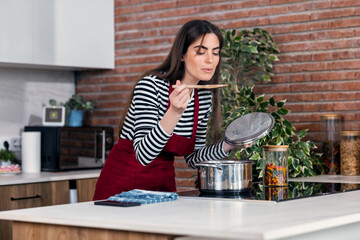 Beautiful woman cooking healthy food in casserole while blowing the spoon to taste the food in the...