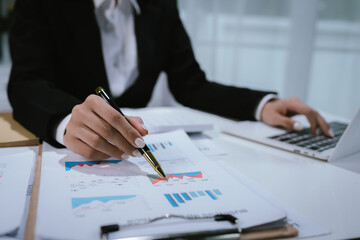A professional businesswoman calculates financial data using a calculator, surrounded by charts and...